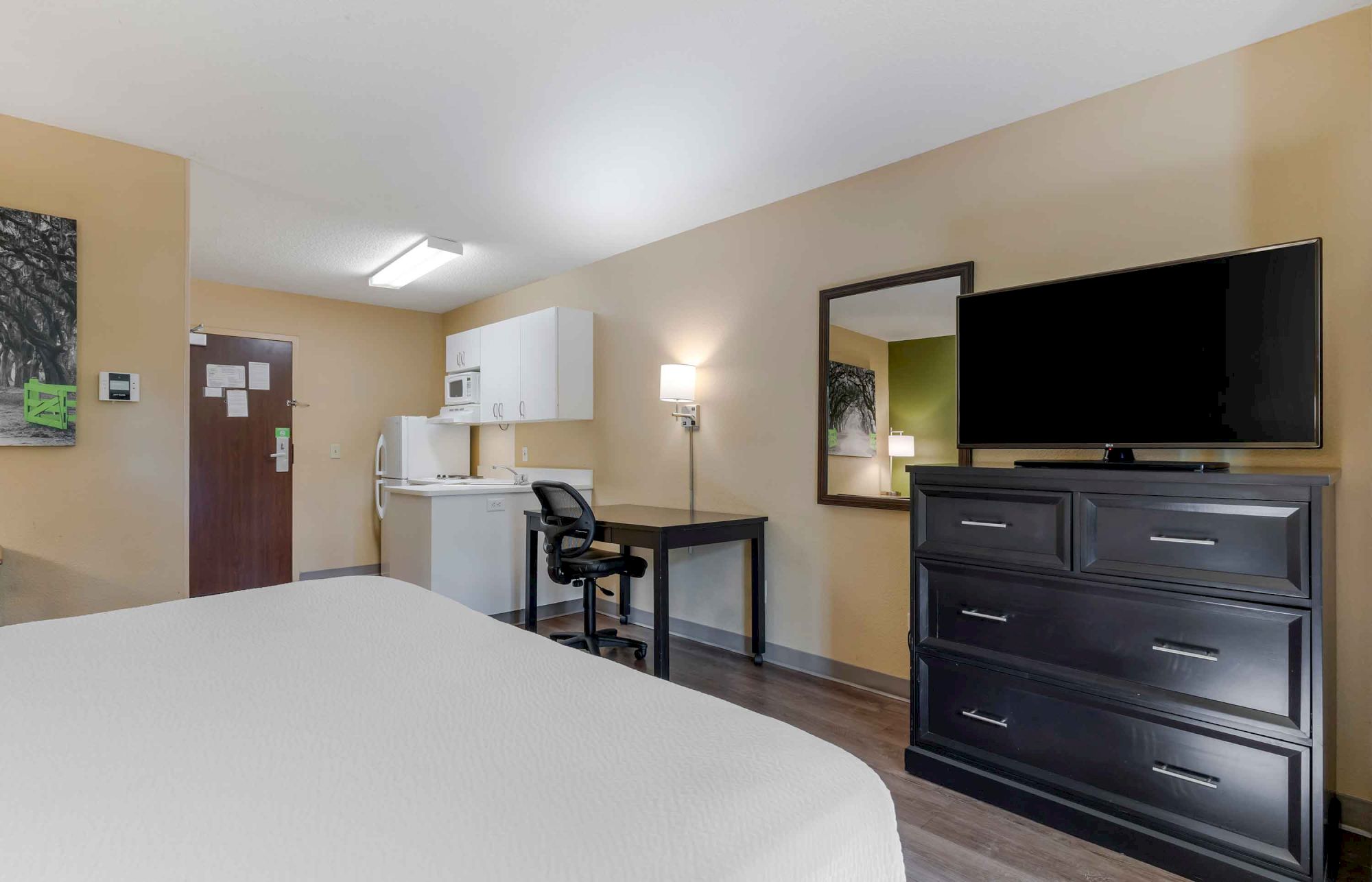 The image shows a hotel room with a bed, TV on a dresser, kitchenette, desk with chair, and wall-mounted mirror.