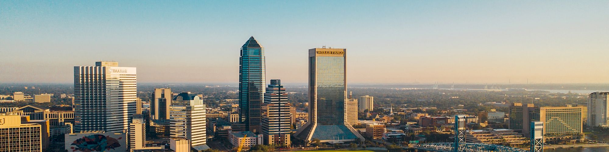 A city skyline featuring tall buildings and a river with a bridge, under a clear blue sky spanning the horizon.