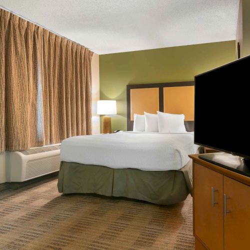 The image shows a hotel room with a queen bed, TV on a cabinet, bedside lamp, brown curtains, and an air conditioning unit.