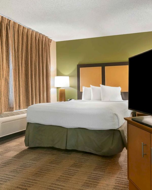 The image shows a hotel room with a queen bed, TV on a cabinet, bedside lamp, brown curtains, and an air conditioning unit.