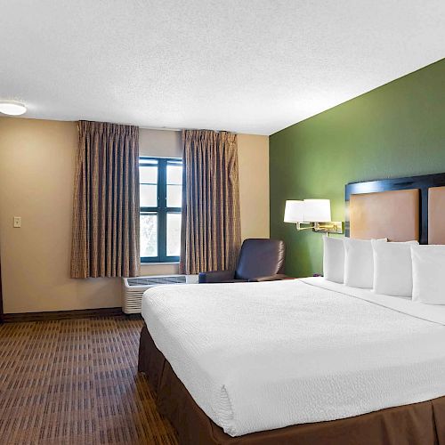 The image shows a hotel room with a neatly made bed, a TV, a window, a nightstand, a chair, a lamp, and a green accent wall.