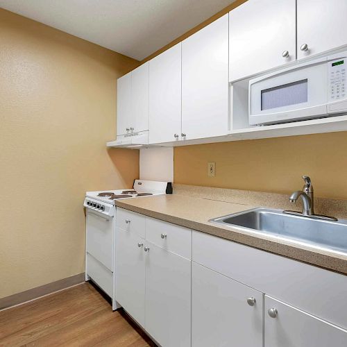 A small kitchen with white cabinets, a microwave, stove, sink, and a refrigerator, alongside a chair and wooden flooring.