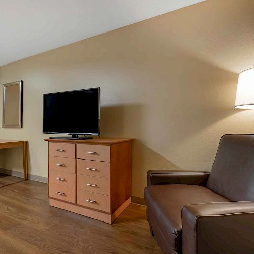 The image shows an interior space with a dark armchair, wooden chest of drawers, TV, desk with a chair, mirror, and a lit lamp on a tan wall.