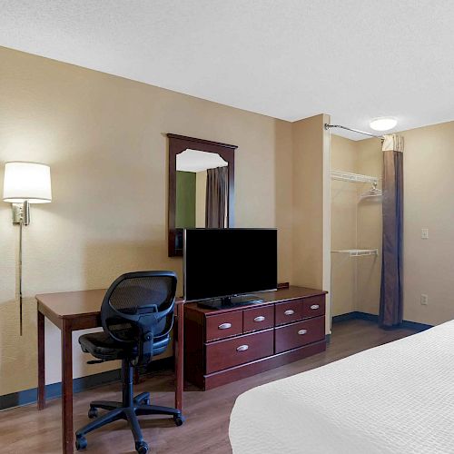 The image shows a hotel room with a bed, desk, chair, TV on a dresser, mirror, and a lamp on the wall, with a closet in the background.