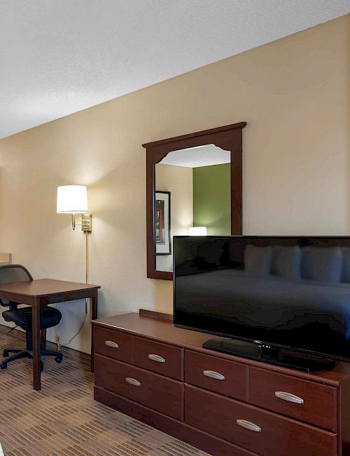 The image shows a modern hotel room with a bed, TV, dresser, desk, chair, and a kitchenette with a microwave and refrigerator.