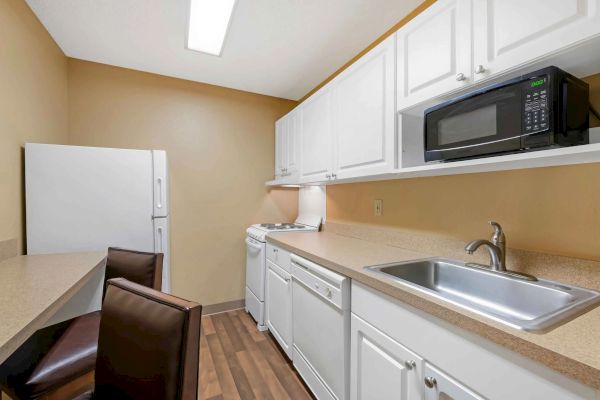 A small, modern kitchen with a white refrigerator, microwave, stove, sink, and brown chairs at a counter.