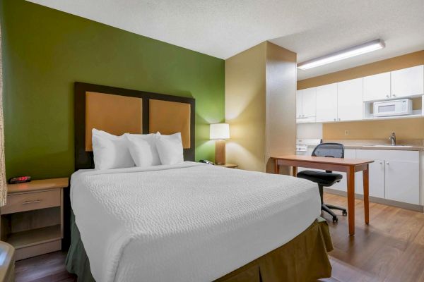 The image shows a hotel room with a double bed, a side table with a lamp, a desk and chair, and a kitchenette with white cabinets and appliances.