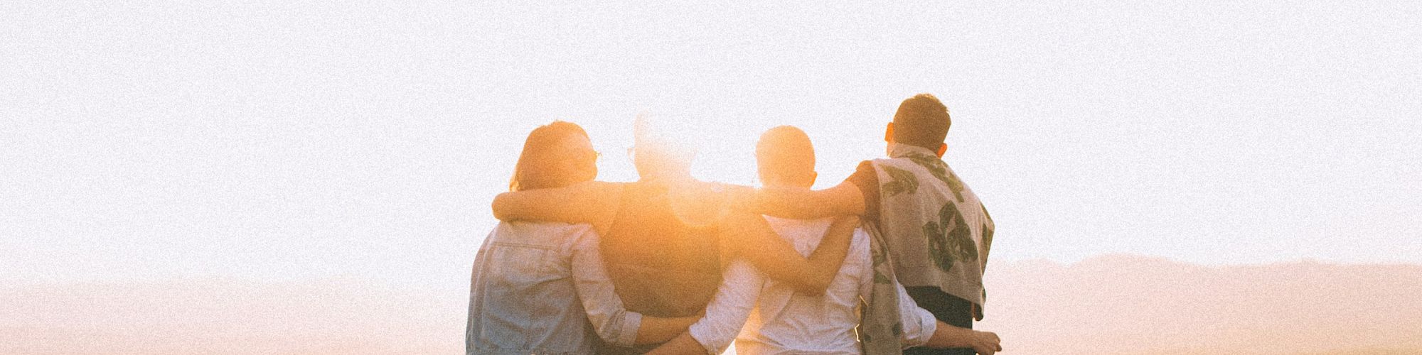 Four people stand arm-in-arm, facing a sunset on a mountaintop, creating a silhouette against the warm light while enjoying the scenic view.