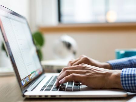 A person in a blue checkered shirt is typing on a laptop, with a wooden desk and window in the background.