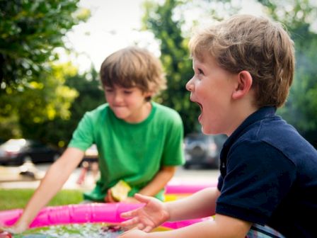 Two children play excitedly outdoors with a small inflatable pool. The older one in green is focused while the younger one in navy beams widely.