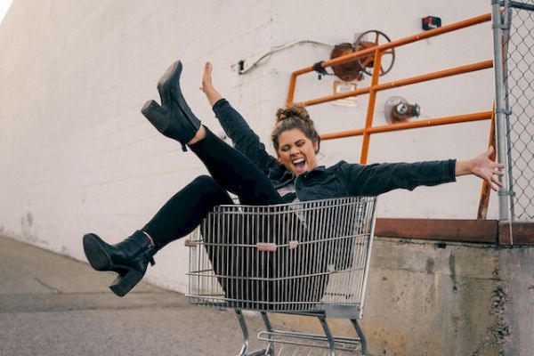 A person is sitting in a shopping cart with their legs up, looking happy near a white wall and orange railing in an outdoor setting.