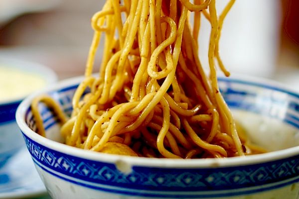 A bowl of noodles held by chopsticks, being lifted from a blue and white patterned bowl, with a blurred background.