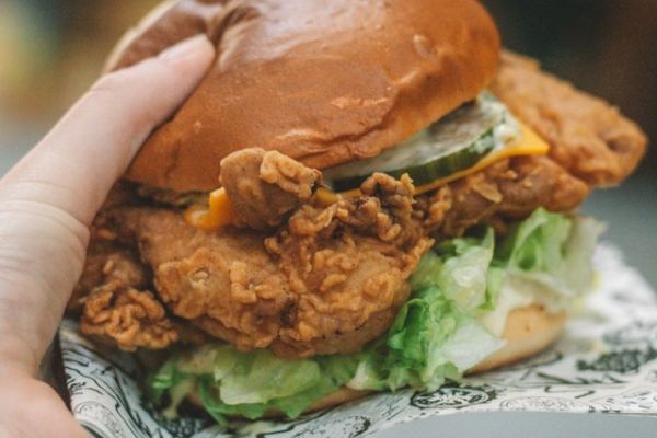 A person is holding a crispy chicken sandwich with lettuce, pickles, and sauce in a brioche bun, wrapped partially in paper.