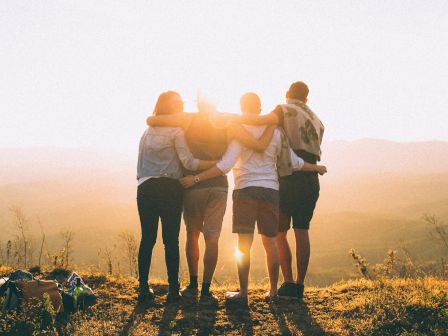 Four people are standing arm-in-arm on a hilltop, watching a scenic sunset together. The atmosphere is warm and serene, with a picturesque view.