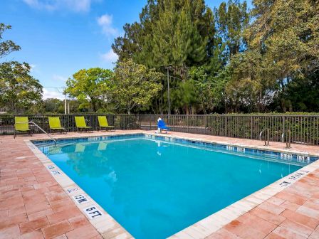 A clean outdoor swimming pool surrounded by a brick deck with green lounge chairs and lush trees in the background, creating a serene setting.