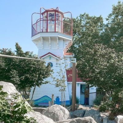 A small, white lighthouse with red railings is surrounded by trees and rocks under a clear blue sky.