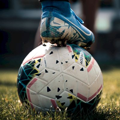 A person wearing blue soccer cleats has one foot resting on a soccer ball on a grassy field, with blurred background.