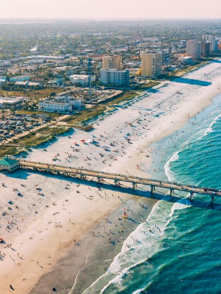 Aerial view of a beach with a pier, people sunbathing and swimming, a coastal city, buildings, and the ocean stretching into the horizon.