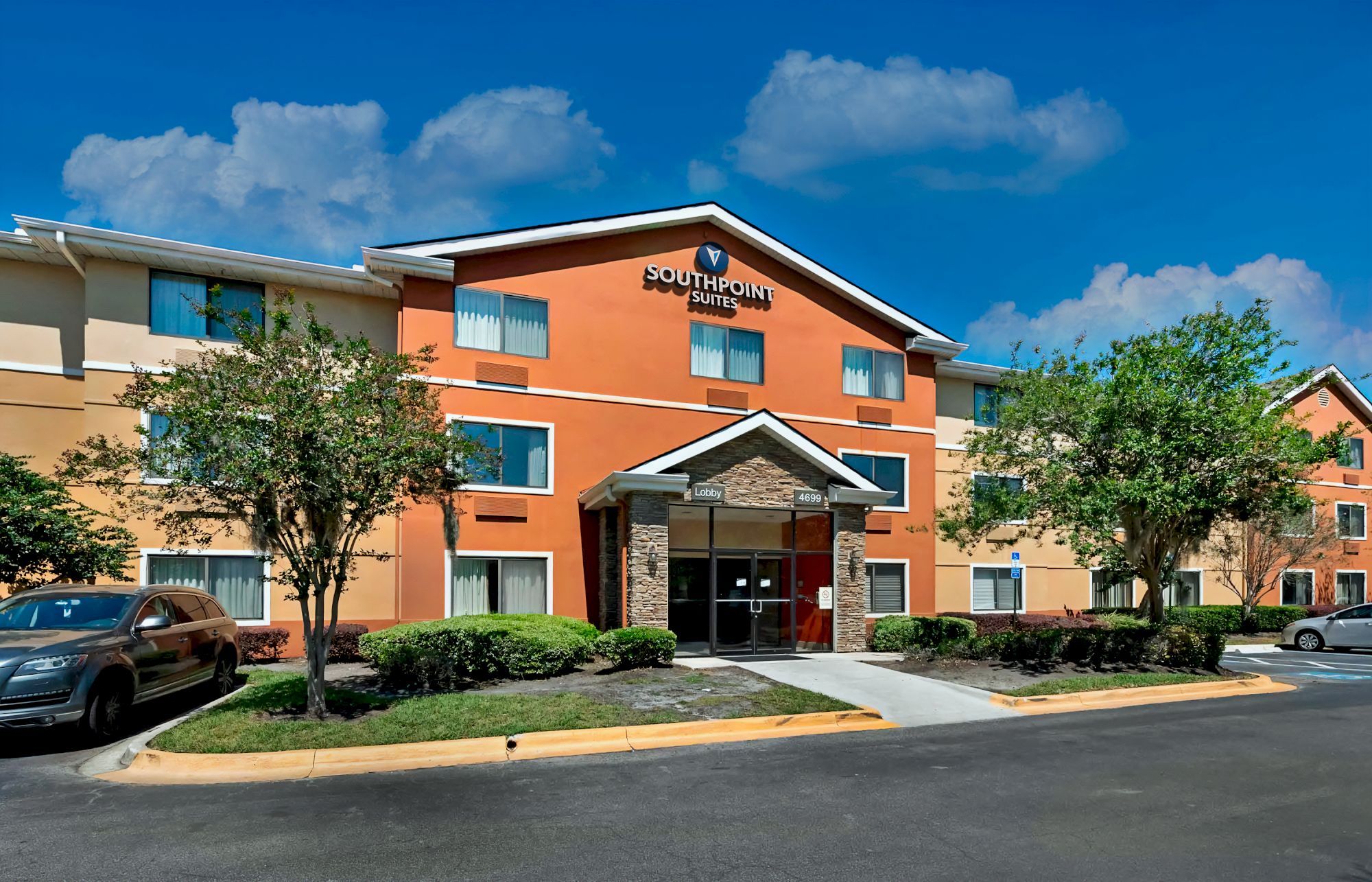 This image shows the exterior of a Southpoint Suites hotel building with a clear blue sky and some cars parked on the side.